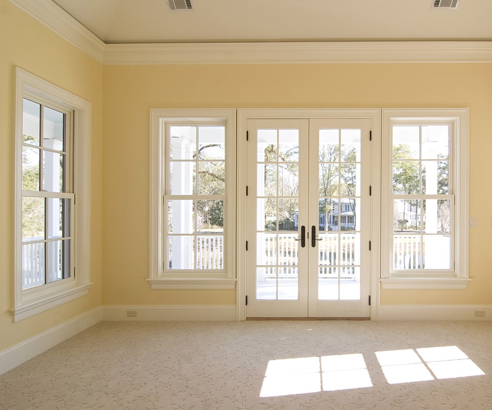 A room with yellow walls, white trim and neutral carpet. Sunlight streams in through energy-efficient custom windows and a set of French-style patio doors.