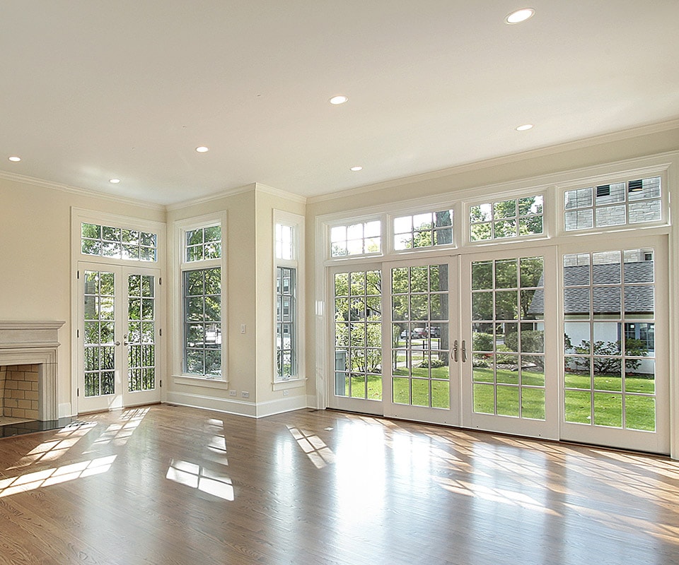 A sunlit room with tan walls, a fireplace and luxurious wood floors. Light streams in through floor-to-ceiling energy-efficient custom windows, and two sets of French-style patio doors.