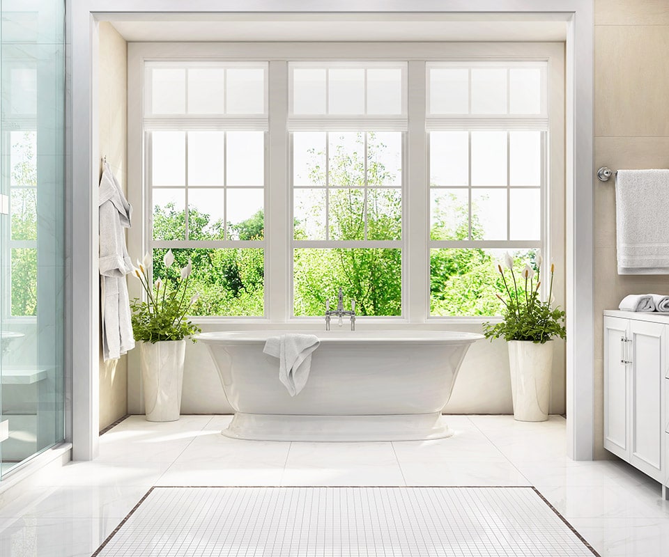 A bright updated bathroom. There is a large bank of windows with landscaping outside. We see an elegant glass shower door to the left. A large, deep, white soaking tub is at center, right by the window, and plants in large white pots flank it. The floor is laid out in premium white tile and mosaic, the walls are a neutral tan, and a whit vanity with chrome fixtures is visible to the right. Bathroom remodeling can transform your space from bland and dingy to beautiful and functional.