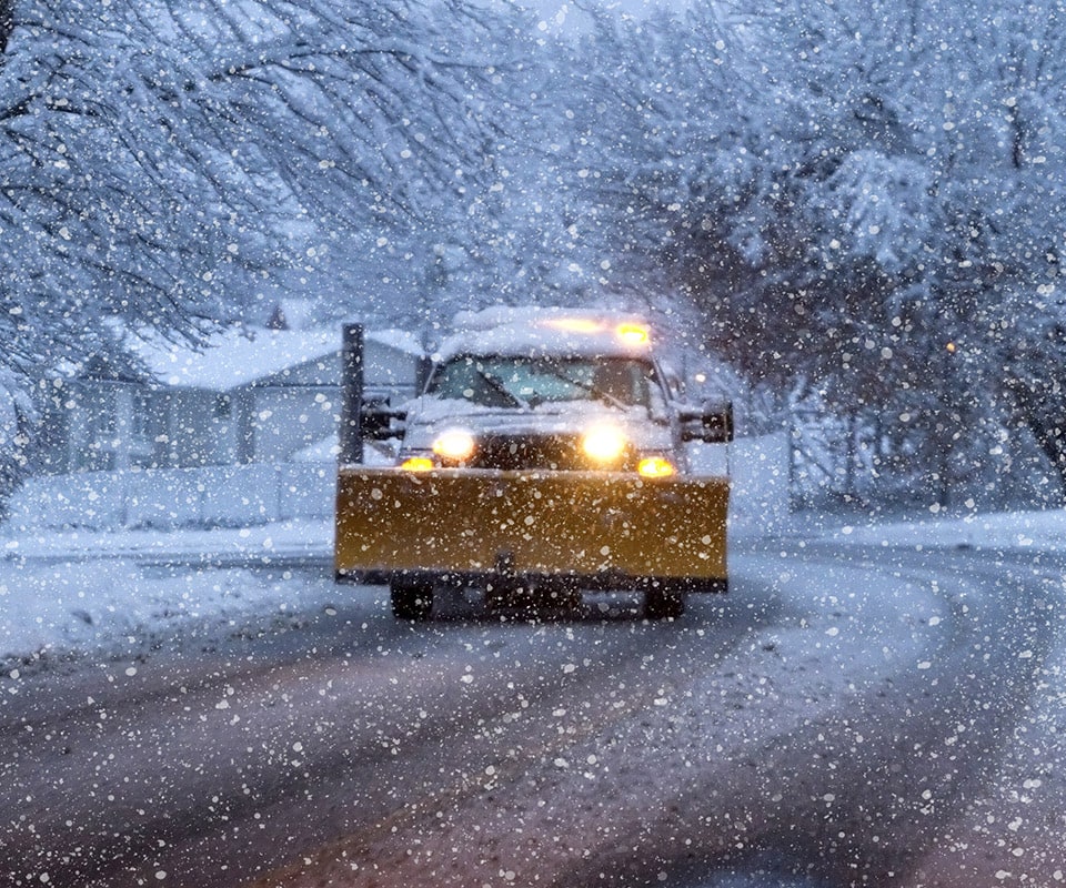 A white truck with a yellow plow travels down the road during a snowstorm. Its lights are on. When you need a snow removal service, give us a call for reliable, prompt service.